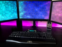 Computer desk setup with 3 monitors and LED strip lights behind it.