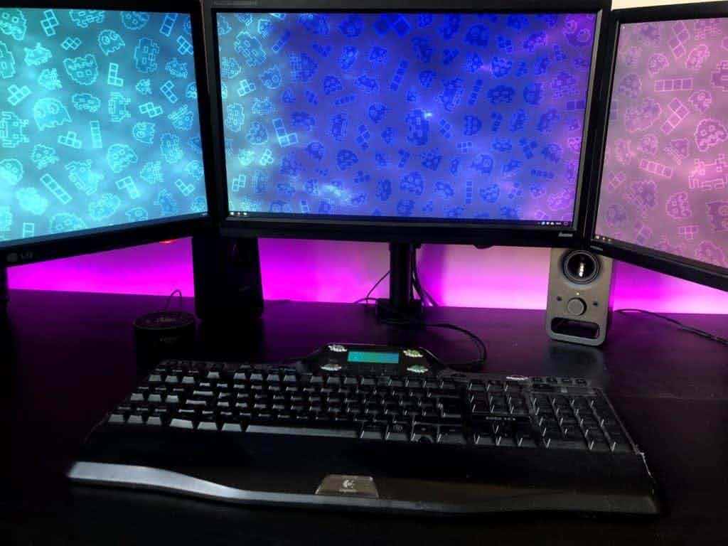 Computer desk setup with 3 monitors and LED strip lights behind it.