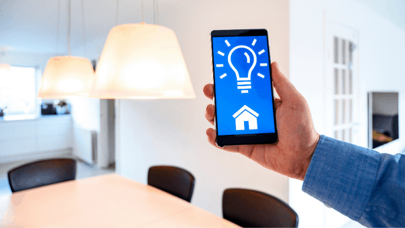 Hand holding smartphone with light bulb on screen, next to a lamp.