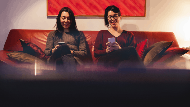 Two girls happy on their smartphones on a red couch in front of a TV.