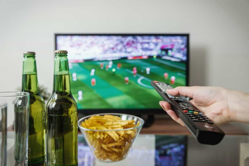 Hand pointing remote at TV with beer and chips nearby