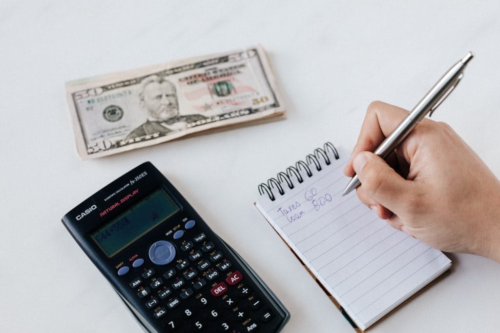 person writing on a notepad with a calculator and a $50 bill