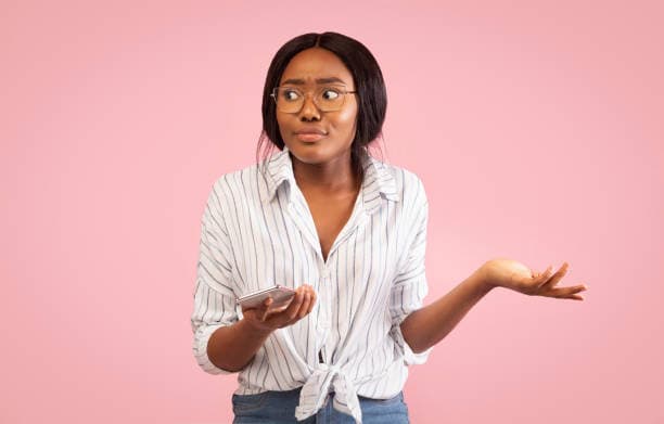 Irritated African American Girl Holding Smartphone And Shrugging Shoulders Looking Aside Standing On Pink Background. Studio Shot